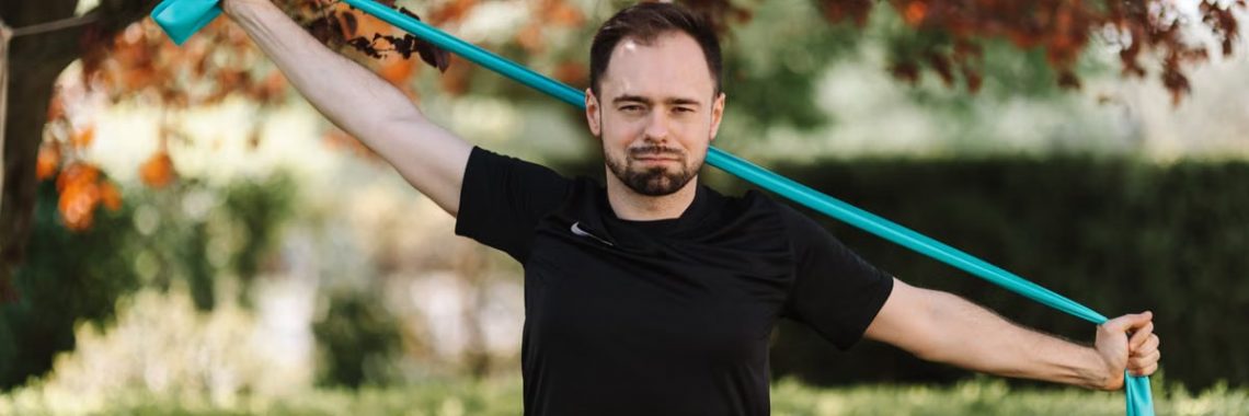 Adult man in black shirt exercises with a resistance band outside.
